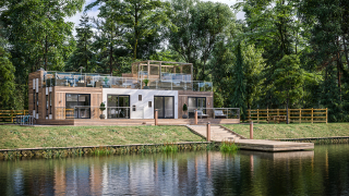 Architectural CGI - modern two-story house with wooden and white facades, extensive glass windows, and multiple balconies overlooks a calm pond. There is a wooden pier extending into the water and lush green trees in the background.
